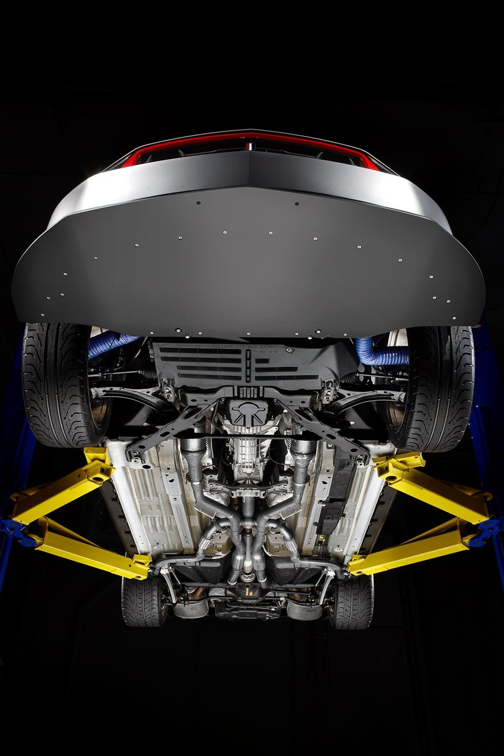This official factory/archive photograph looks straight up at the underside of a 2012 Ford Mustang Boss 302, showing the flat belly pan, front subframe, driveshaft and the quad exhaust with its side outlets.