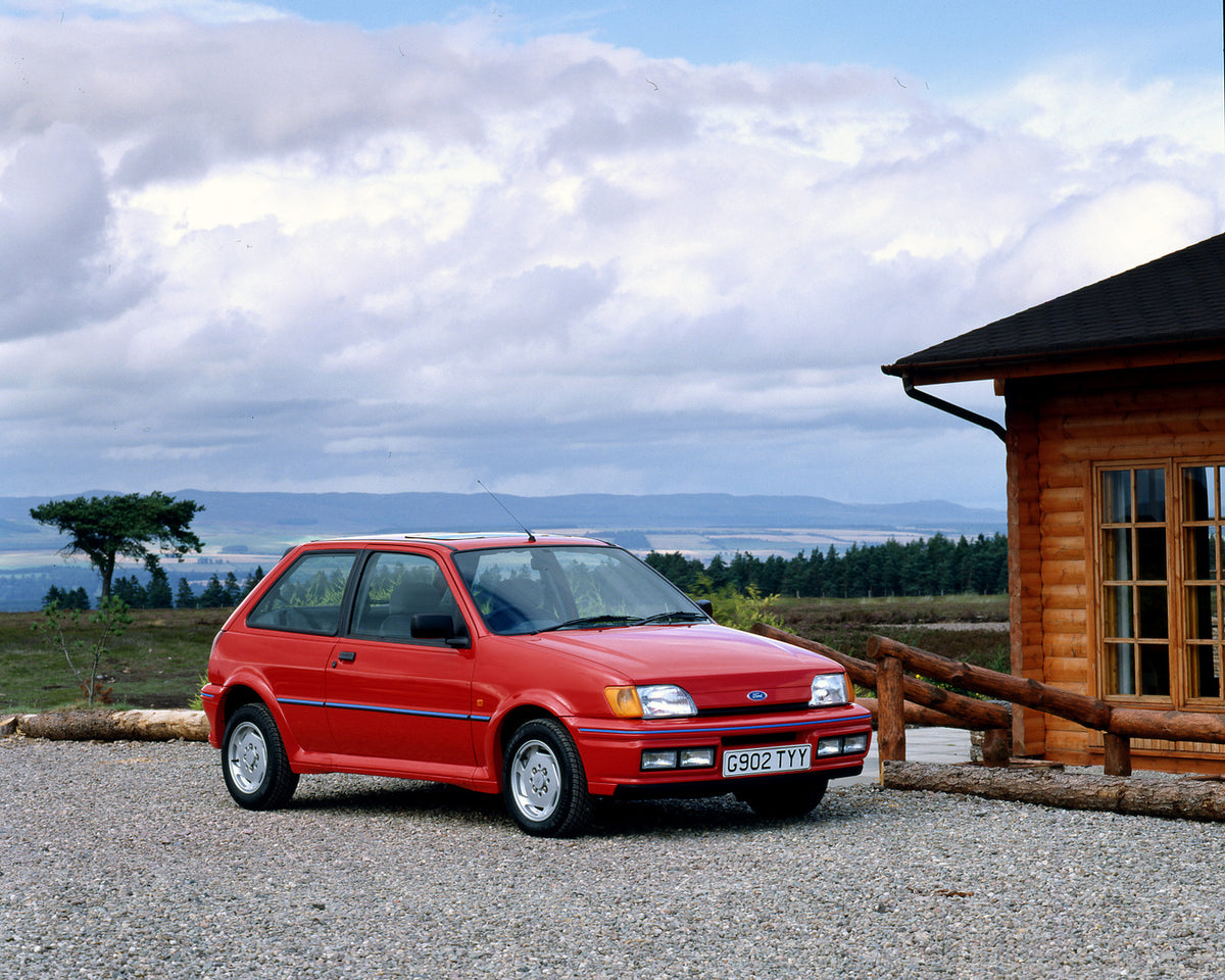 This archival image documents the 1989 Ford Fiesta Mark III XR2i in driveway of cabin with scenic trees and mountains and is preserved in the Ford Heritage Vault as part of Ford Motor Company's historic photographic archive. Free for personal, non...