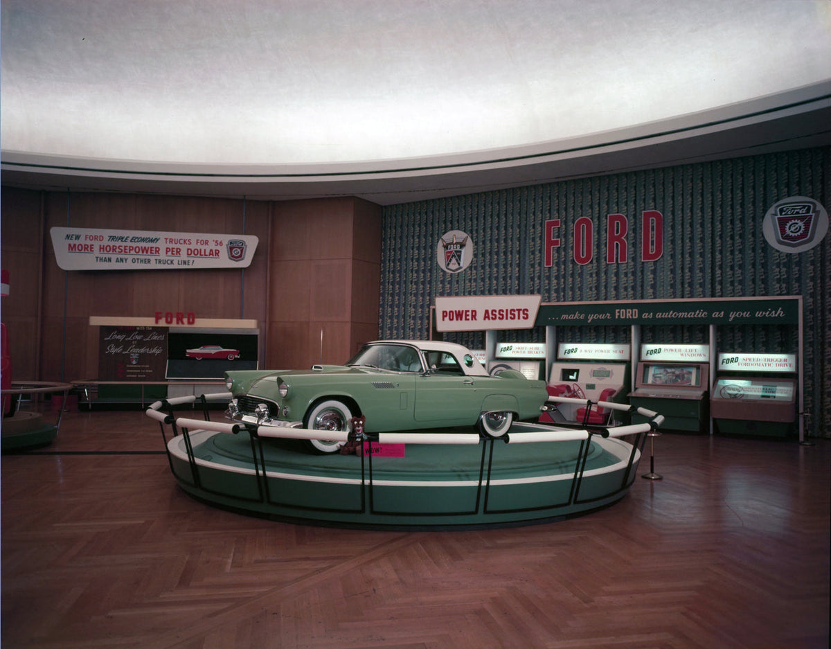 A green 1956 Ford Thunderbird with a light-colored hardtop stands on a circular display platform in a showroom.