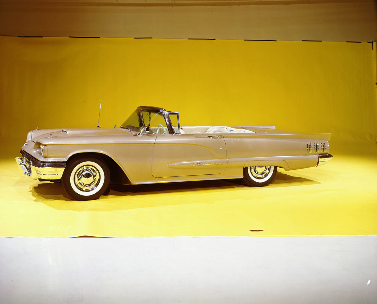 A light-colored 1960 Ford Thunderbird convertible is shown against a yellow studio backdrop.