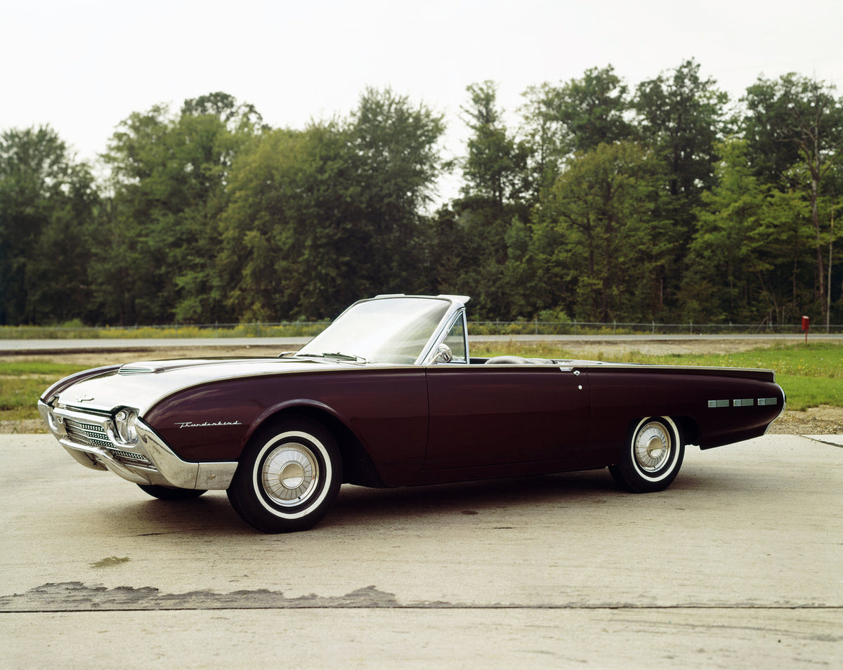 A dark 1962 Ford Thunderbird convertible is photographed on a paved turnout with trees behind it.