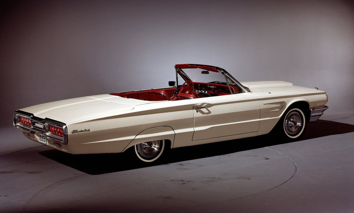 A white 1965 Ford Thunderbird convertible is photographed from the rear against a dark studio background.