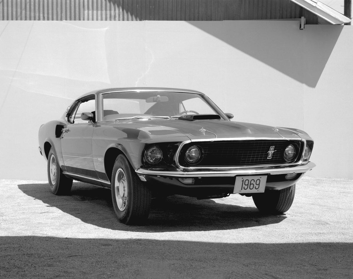 A 1969 Ford Mustang Mach 1 is photographed from the front three-quarter angle outside a building in black and white.