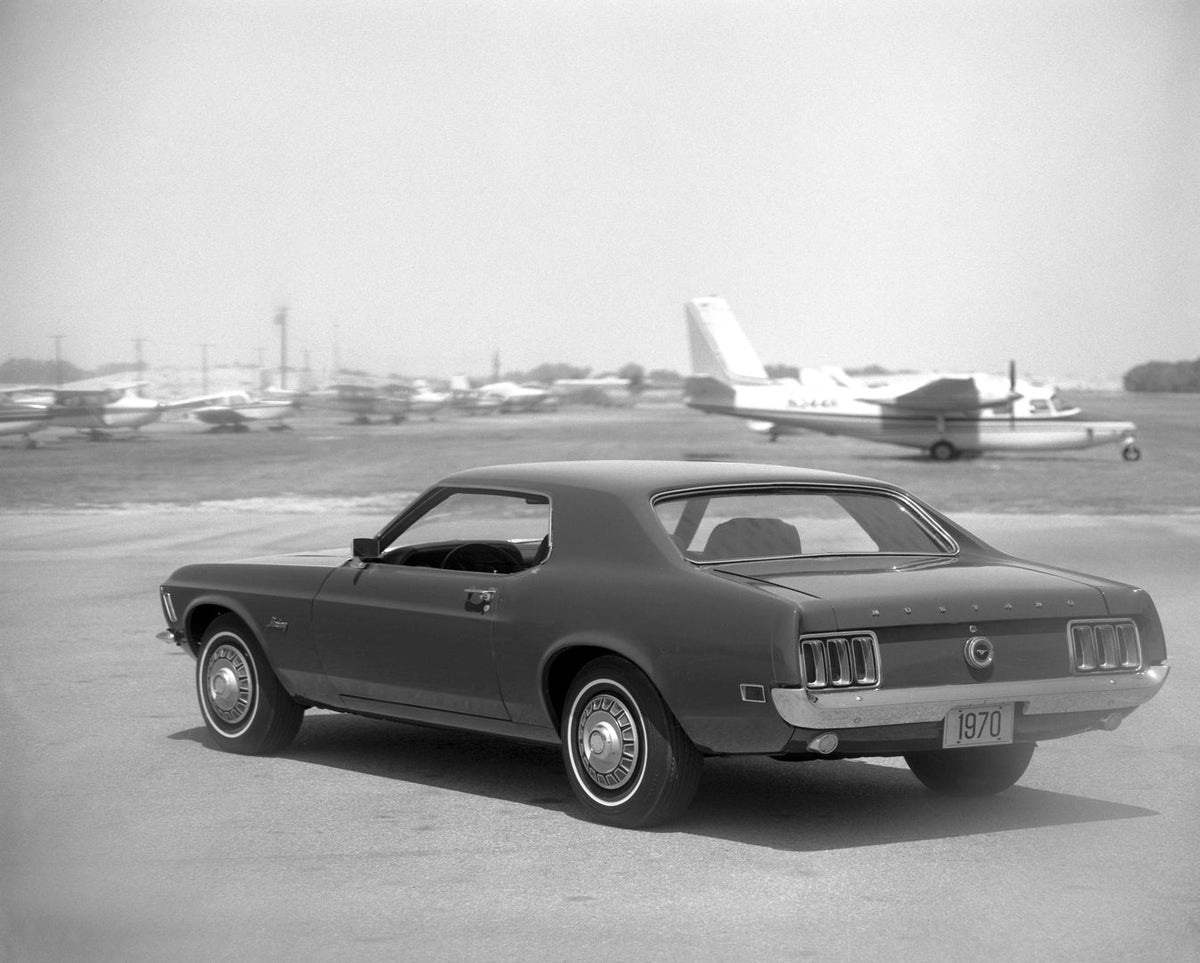 A 1970 Ford Mustang hardtop is photographed from the rear three-quarter angle at an airport in black and white.
