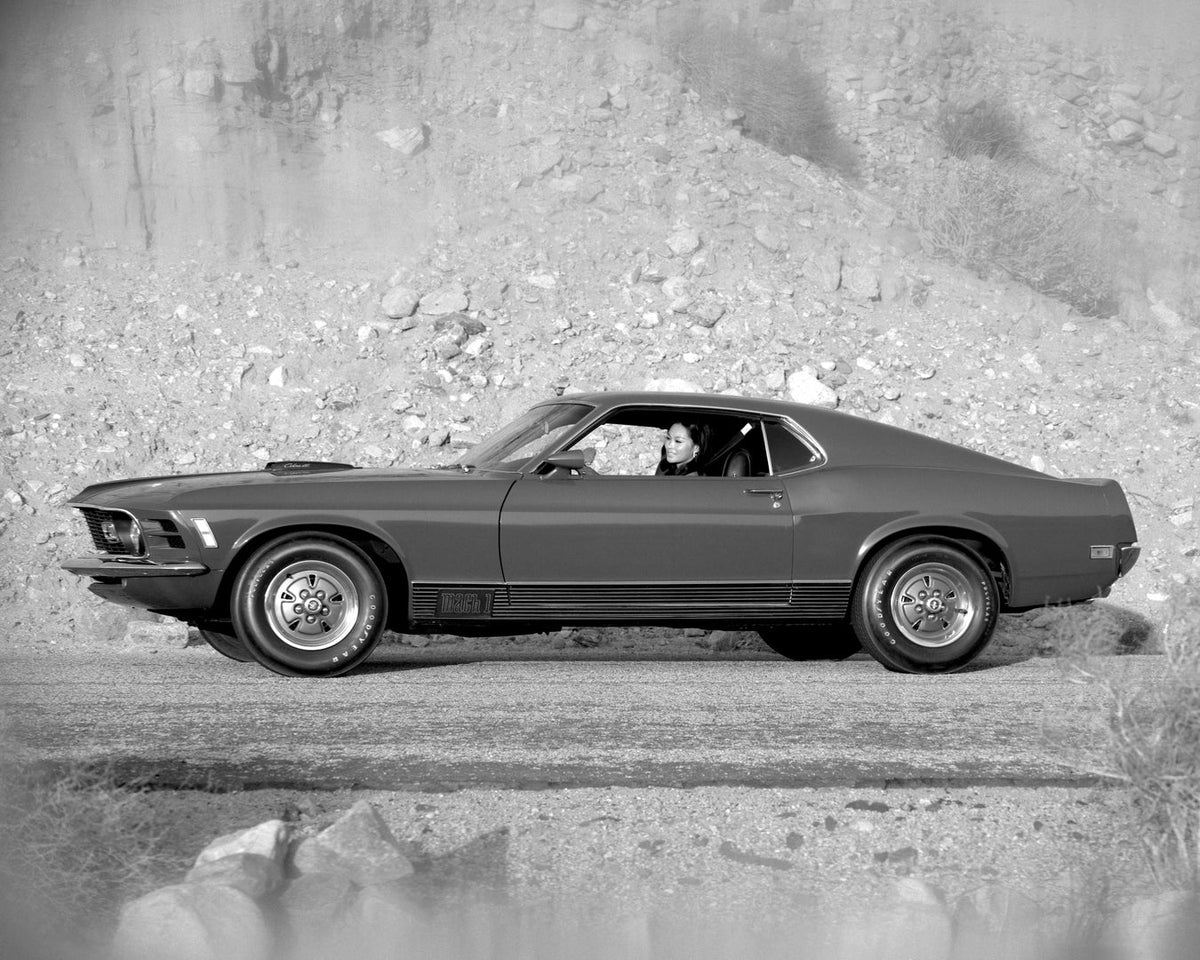A 1970 Ford Mustang Mach 1 is photographed in side profile beside a rocky embankment in black and white.