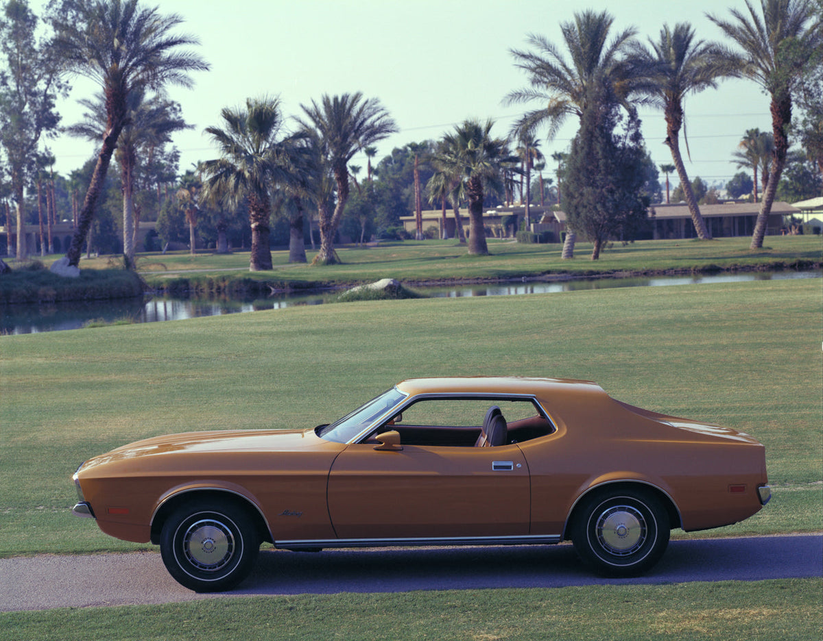 A bronze-colored 1972 Ford Mustang hardtop is parked beside a palm-lined lawn.