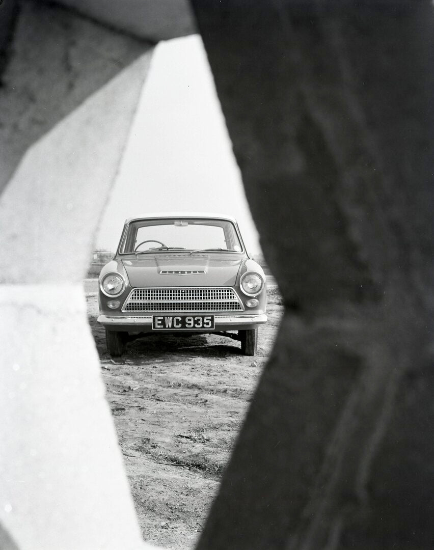 This archival image documents the 1962 Ford Cortina Deluxe Mark I front view through fence and is preserved in the Ford Heritage Vault as part of Ford Motor Company's historic photographic archive. Free for personal, non-commercial use. Ford Herit...